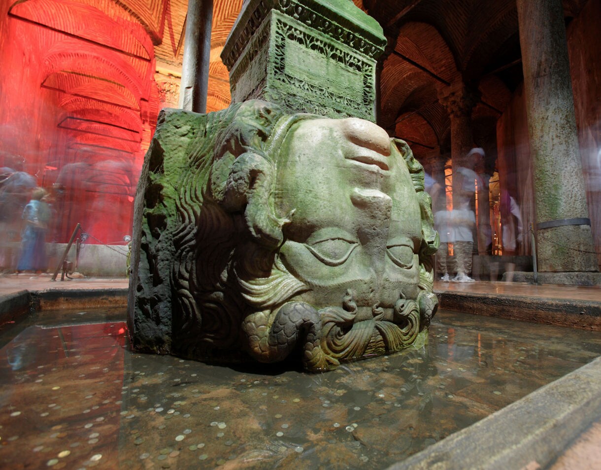 An upside-down Medusa head carved in stone, used as a column base in the dimly lit Basilica Cistern with glowing red arches overhead.